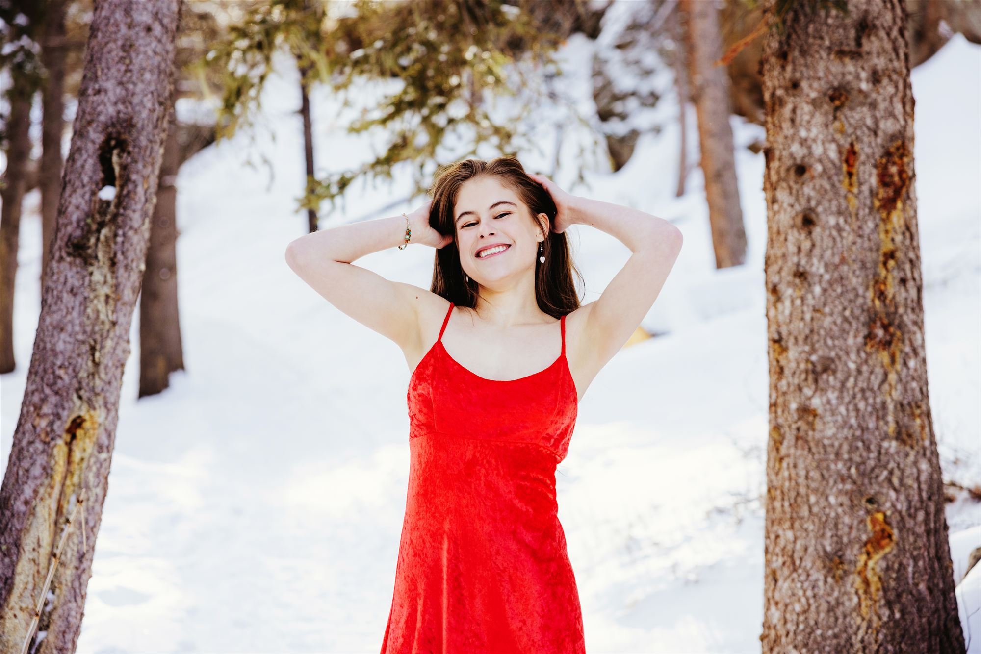 A woman laughing in a red velvet handmade dress, standing among snow-covered pine trees.